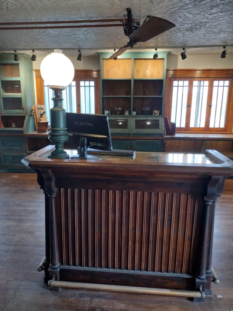 A wooden hotel reception desk with a lamp and computer monitor sits in a rustic room with wooden shelves and large windows in the background, capturing the inviting charm of Chama.